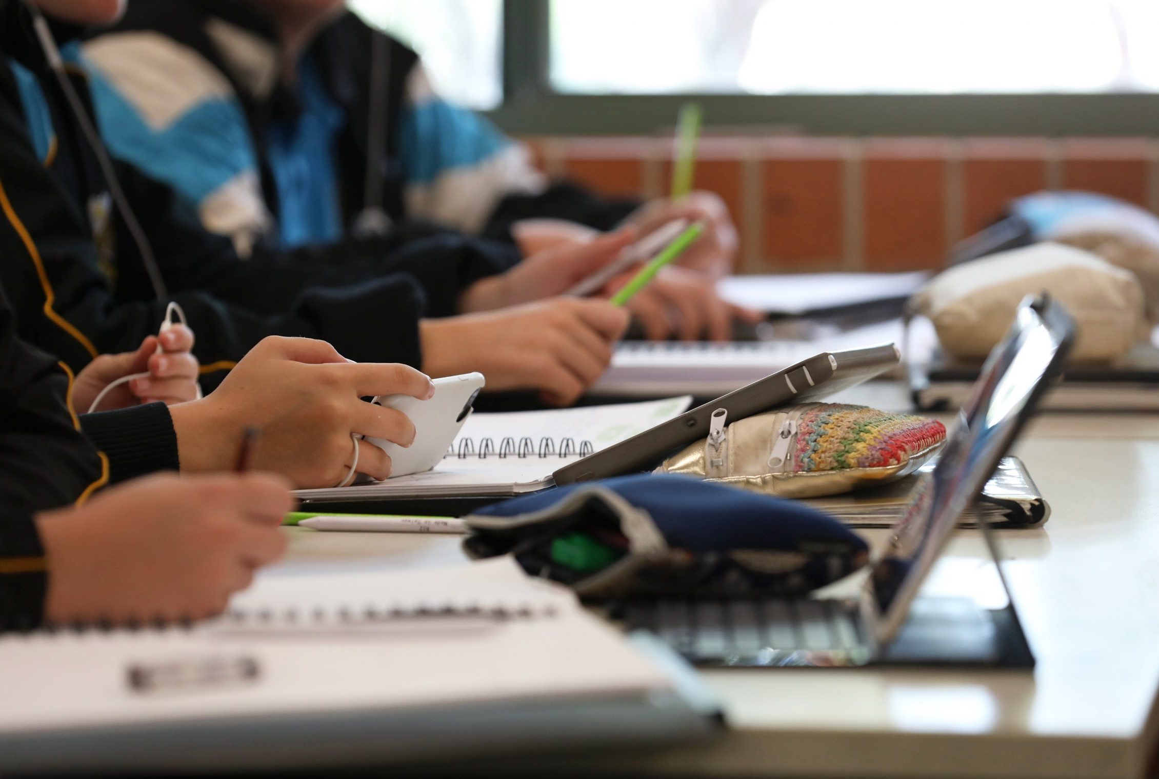A student in uniform distracted holding using and watching a mobile phone during a lesson at high school. Books, tablets and pencil cases all visible on the desk and work space.